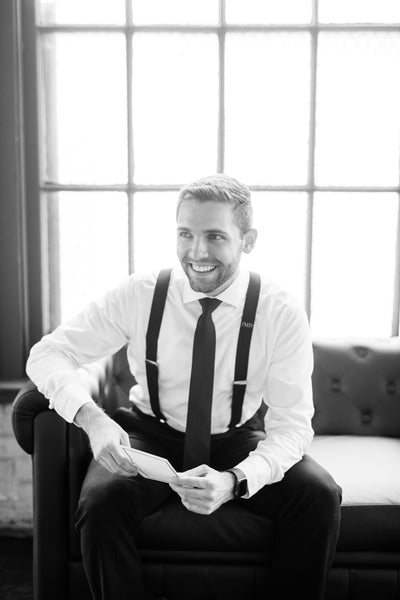 groom reading note from bride for wedding wearing black tie and monogram suspender