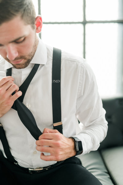 groom tying black bow tie for wedding with monogram suspender