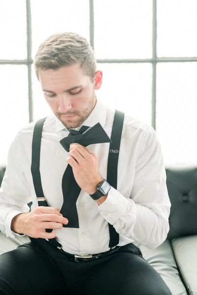 man tying bow tie for black tie wedding wearing monogram suspender